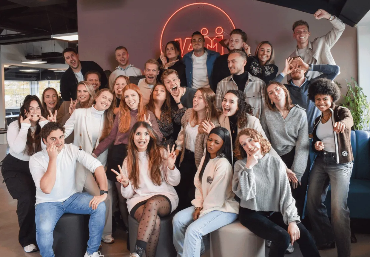 The whole JAM team poses smiling under a red neon logo in the office