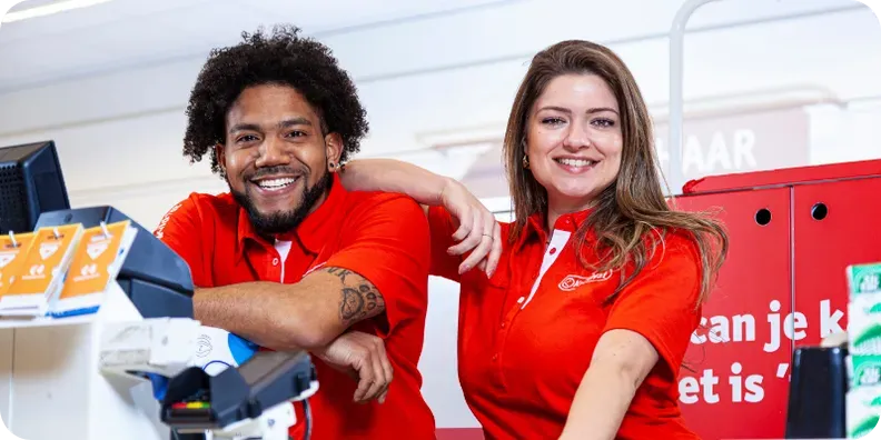 Two smiling Kruidvat employees in red polo shirts behind the till of a store