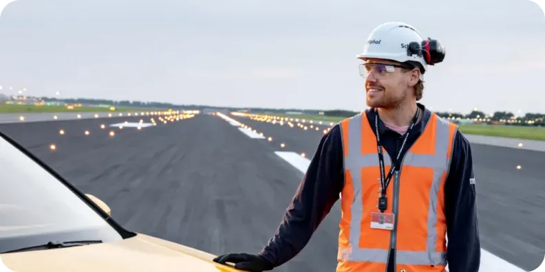 Schiphol employee in a helmet and orange high-visibility vest beside a vehicle on the runway